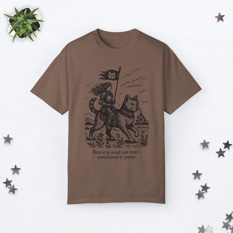Peut inclure: Un t-shirt marron avec une illustration en noir et blanc d'une femme chevauchant un chat. Le chat porte un collier et la femme tient un drapeau. Le texte "Bravery need not roar - sometimes it purrs." est imprim&eacute; sous l'illustration.