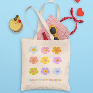 Pu&ograve; includere: Una borsa tote in tela bianca con un design di fiori colorati con facce sorridenti. Il testo "think happy thoughts" &egrave; stampato sul fondo della borsa.
