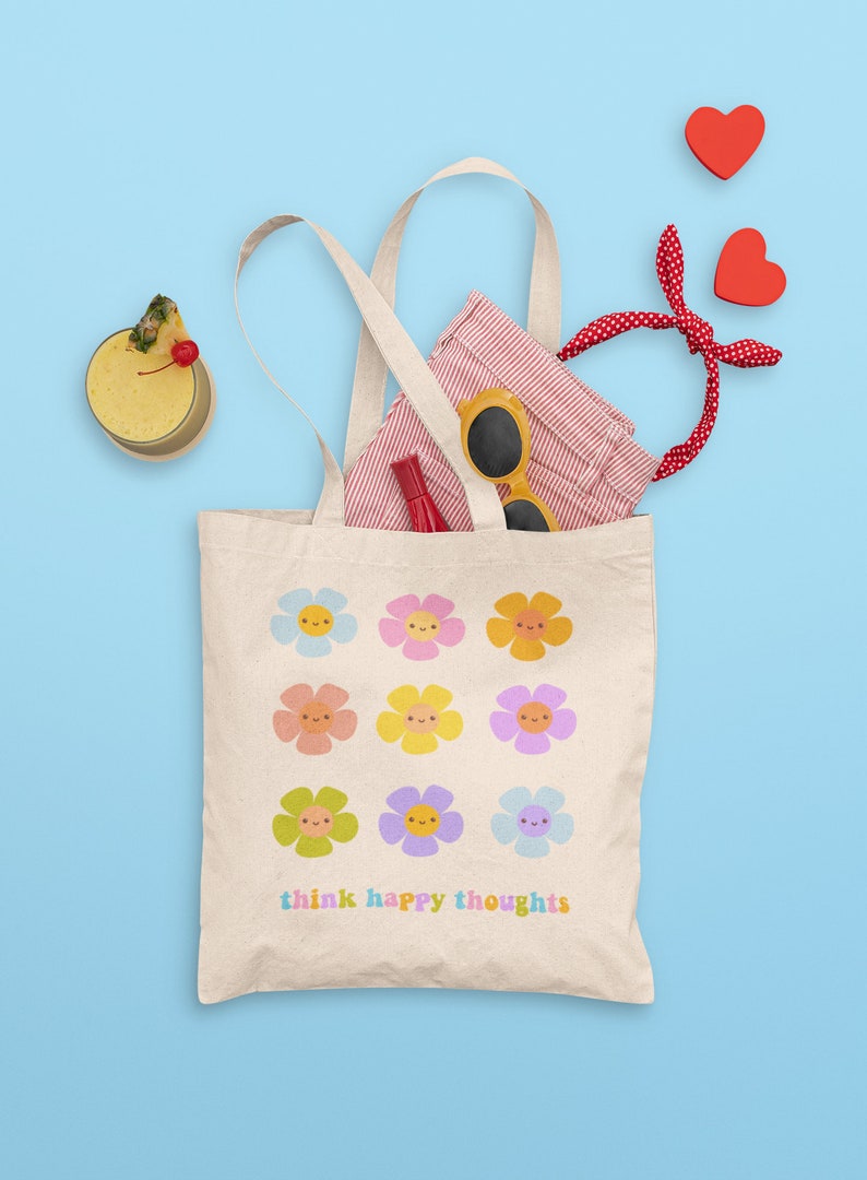 Pu&ograve; includere: Una borsa tote in tela bianca con un design di fiori colorati con facce sorridenti. Il testo "think happy thoughts" &egrave; stampato sul fondo della borsa.