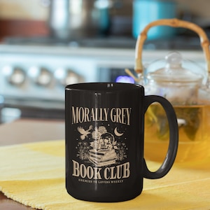 May include: Black ceramic mug with a white design. The design features a skull with a book on top, a bird flying above, and the text "Morally Grey Book Club Enemies to Lovers Weekly".