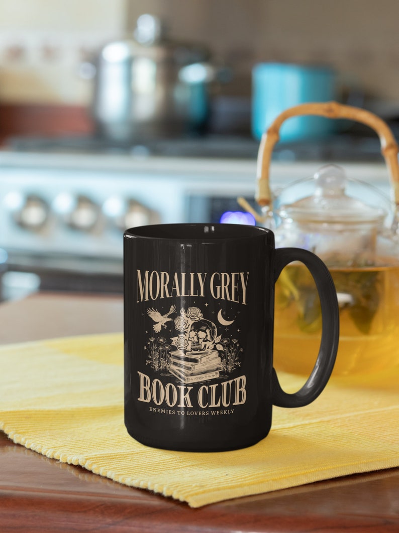 May include: Black ceramic mug with a white design. The design features a skull with a book on top, a bird flying above, and the text "Morally Grey Book Club Enemies to Lovers Weekly".