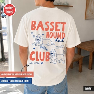 May include: An ivory short-sleeve t-shirt featuring a graphic on the back with the text "BASSET HOUND CLUB" in large coral letters. The design includes two blue line-art illustrations of basset hounds, paw prints, a heart, and "est. 2026".