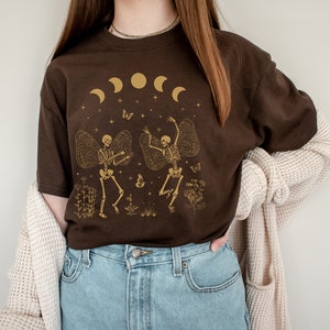 May include: A brown t-shirt with a graphic of two skeletons dancing with wings. The skeletons are gold and the background is a dark brown with a crescent moon and stars.