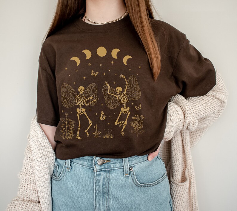 May include: A brown t-shirt with a graphic of two skeletons dancing with wings. The skeletons are gold and the background is a dark brown with a crescent moon and stars.
