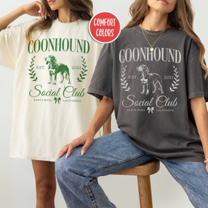 May include: Two women wearing oversized t-shirts with a graphic design of a coonhound dog and the text "COONHOUND Social Club EST. 2021 SANTA ROSA CALIFORNIA". The t-shirt on the left is white and the t-shirt on the right is charcoal grey.