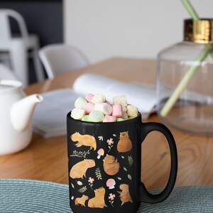 May include: Black ceramic mug with a white handle. The mug features a colorful illustration of capybaras, flowers, and mushrooms. The mug is filled with pastel-colored marshmallows.