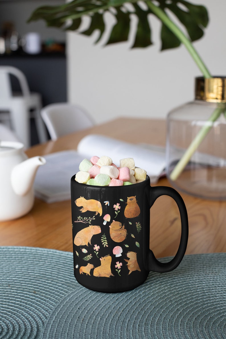 May include: Black ceramic mug with a white handle. The mug features a colorful illustration of capybaras, flowers, and mushrooms. The mug is filled with pastel-colored marshmallows.