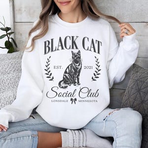 May include: White sweatshirt with a black cat graphic and the text "Black Cat Social Club Est. 2021 Lonsdale Minnesota".