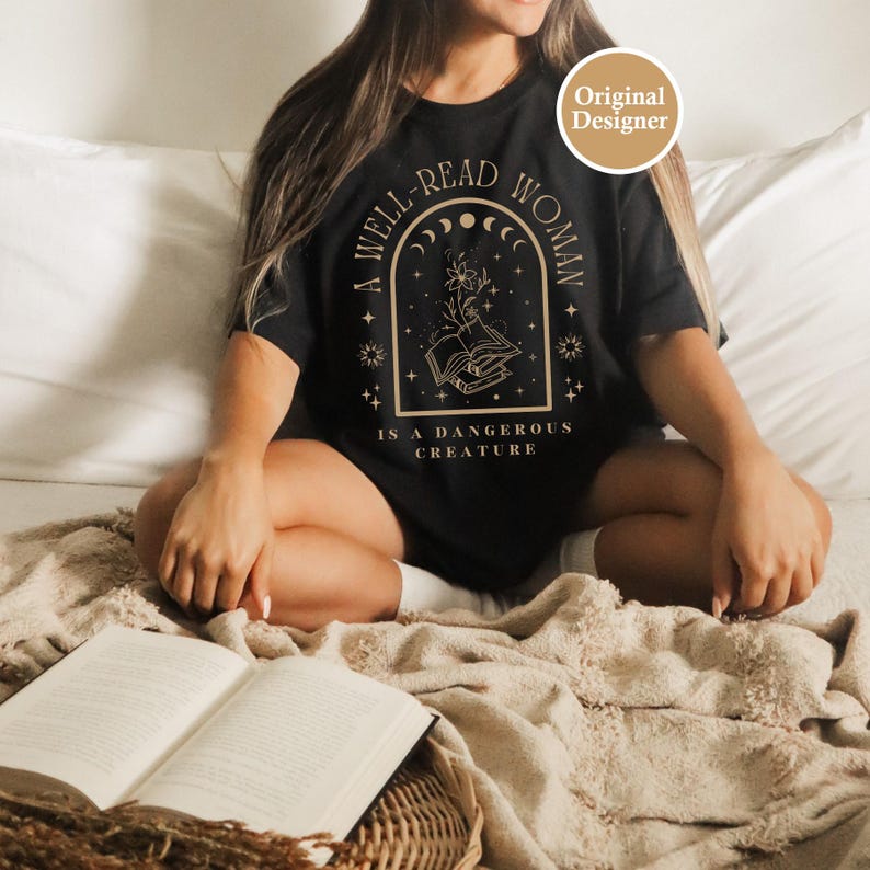 K&ouml;nnte beinhalten: Schwarzes T-Shirt mit dem goldenen Schriftzug "A WELL-READ WOMAN IS A DANGEROUS CREATURE". Das Design zeigt ein offenes Buch, Blumen und Himmelselemente. Das Shirt ist ein Freizeitartikel.