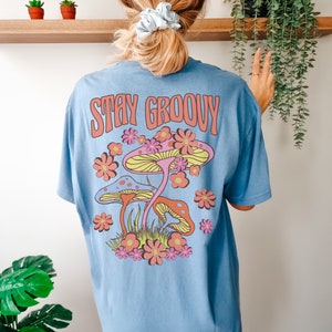 May include: A light blue t-shirt with a graphic of colorful mushrooms and flowers. The text "Stay Groovy" is printed in pink on the back of the shirt.