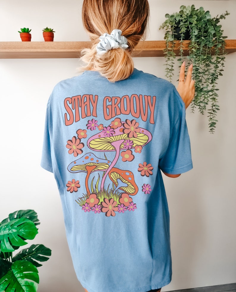 May include: A light blue t-shirt with a graphic of colorful mushrooms and flowers. The text "Stay Groovy" is printed in pink on the back of the shirt.
