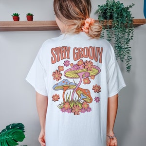 May include: White t-shirt with a graphic of colorful mushrooms and flowers. The text "Stay Groovy" is printed in orange on the back of the shirt.