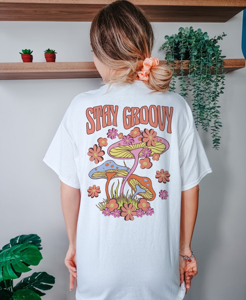 May include: White t-shirt with a graphic of colorful mushrooms and flowers. The text "Stay Groovy" is printed in orange on the back of the shirt.