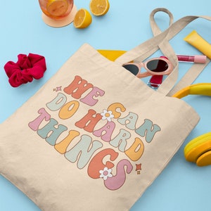 Op de afbeelding: Een beige canvas tote bag met de tekst "We Can Do Hard Things" in regenboogkleuren. De tas is gevuld met verschillende items, waaronder een zonnebril, een haarband en lipgloss.