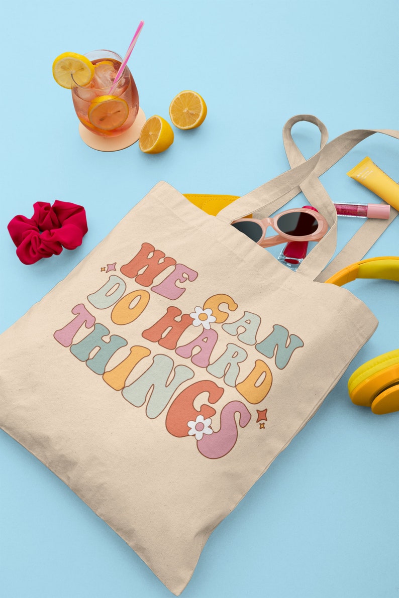 Op de afbeelding: Een beige canvas tote bag met de tekst "We Can Do Hard Things" in regenboogkleuren. De tas is gevuld met verschillende items, waaronder een zonnebril, een haarband en lipgloss.