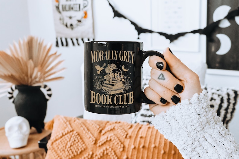 May include: Black coffee mug with a gold design that says "Morally Grey Book Club Enemies to Lovers Weekly".