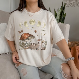 Puede incluir: Una camiseta de color crema con un gr&aacute;fico de un hongo, una familia de zarig&uuml;eyas y una luna creciente con estrellas. El gr&aacute;fico est&aacute; en tonos de marr&oacute;n, verde y amarillo.