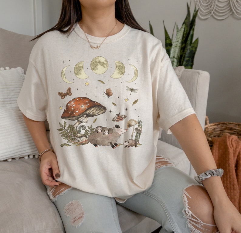 Puede incluir: Una camiseta de color crema con un gr&aacute;fico de un hongo, una familia de zarig&uuml;eyas y una luna creciente con estrellas. El gr&aacute;fico est&aacute; en tonos de marr&oacute;n, verde y amarillo.