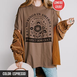 May include: Espresso-colored t-shirt with the text "NO KINGS IN AMERICA SINCE 1776 PROTECT DEMOCRACY" in black. The design includes a floral arrangement, sun, and moon. The shirt is part of the Comfort Colors collection.