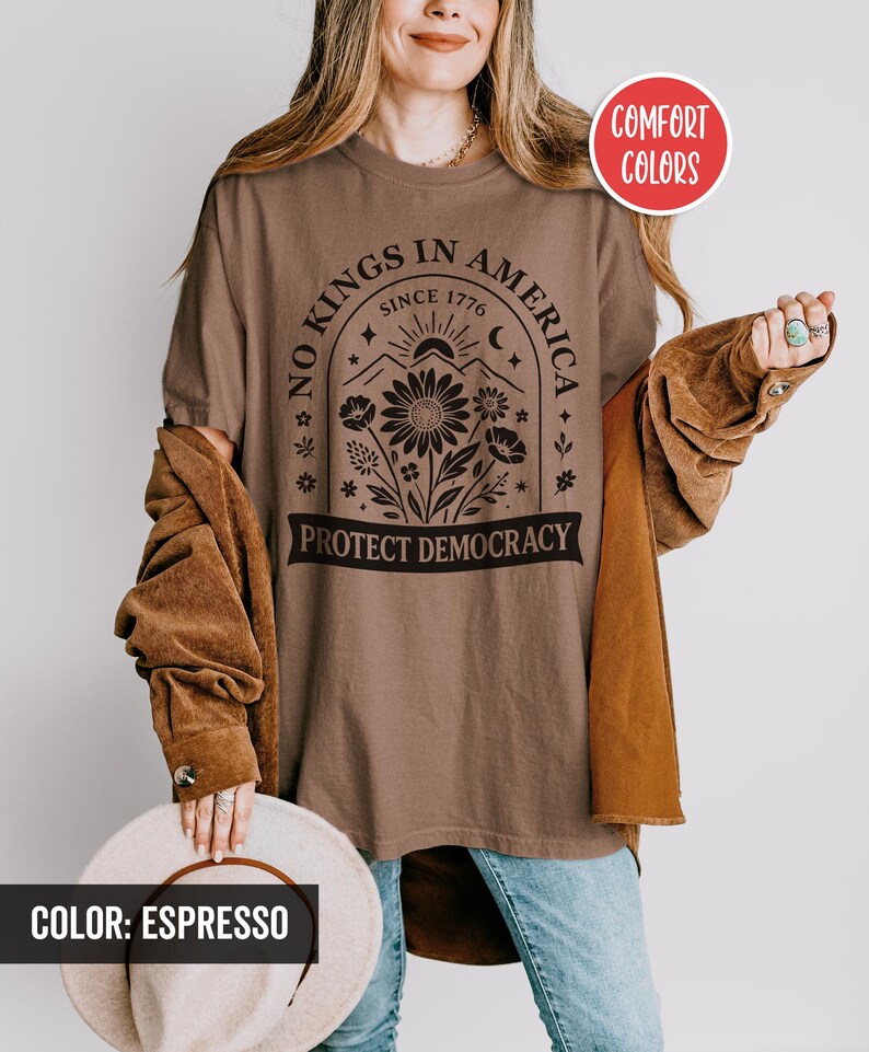 May include: Espresso-colored t-shirt with the text "NO KINGS IN AMERICA SINCE 1776 PROTECT DEMOCRACY" in black. The design includes a floral arrangement, sun, and moon. The shirt is part of the Comfort Colors collection.