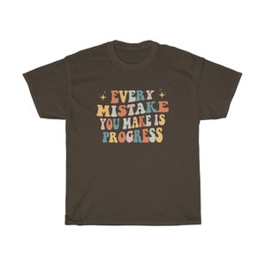 May include: A dark brown t-shirt with the text "EVERY MISTAKE YOU MAKE IS PROGRESS" in colorful, retro-style lettering. The text is in shades of orange, blue, pink, and yellow, with star accents. The shirt is a casual wear item.