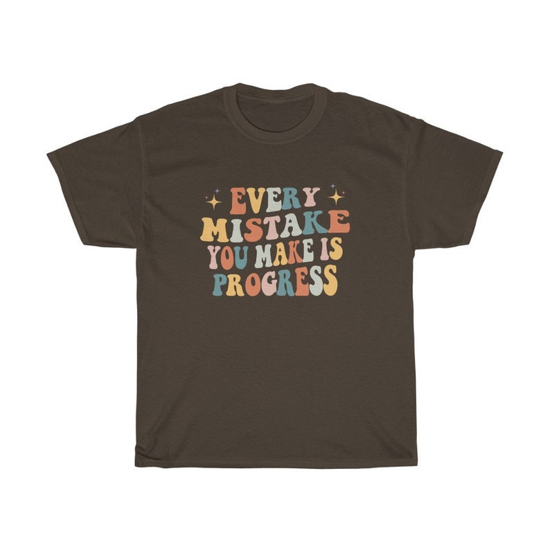 May include: A dark brown t-shirt with the text "EVERY MISTAKE YOU MAKE IS PROGRESS" in colorful, retro-style lettering. The text is in shades of orange, blue, pink, and yellow, with star accents. The shirt is a casual wear item.