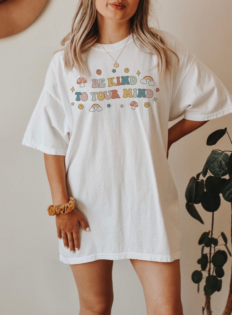 Puede incluir: Camiseta blanca de gran tama&ntilde;o con un estampado gr&aacute;fico del texto "Be Kind To Your Mind" en letras de colores con detalles de arco&iacute;ris, setas y estrellas.