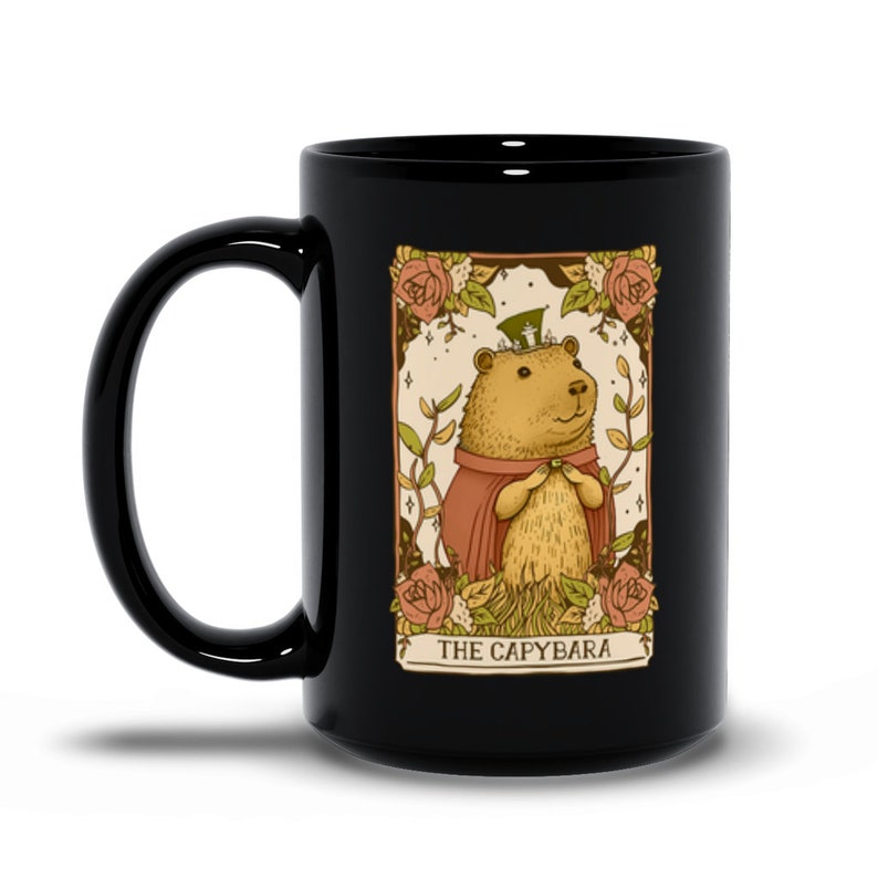 May include: Black ceramic mug with a white illustration of a capybara wearing a green hat and a red robe. The illustration is surrounded by flowers and the text "THE CAPYBARA" is at the bottom.
