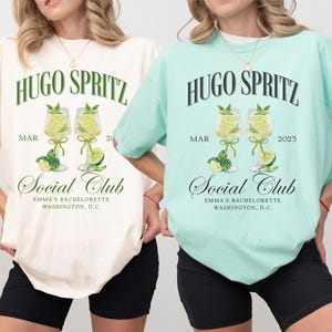 May include: Two white t-shirts with a green and black graphic design. The graphic features the text "HUGO SPRITZ" and two glasses of a lime-colored drink with lime slices. The text "MAR 20" and "MAR 2025" are also included in the design. The text "Social Club" is below the glasses and the text "EMMA'S BACHELORETTE WASHINGTON, D.C." is below that. The t-shirt on the left is ivory and the t-shirt on the right is light blue.