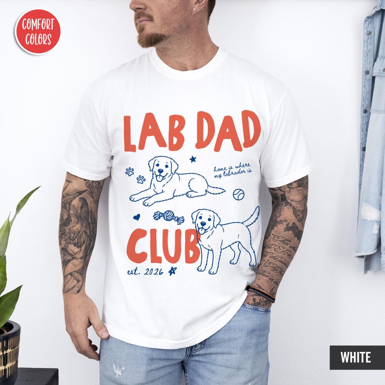 Puede incluir: Una camiseta blanca de manga corta con el texto rojo LAB DAD CLUB y el texto azul est. 2026. El gr&aacute;fico incluye dos perros Labrador en dibujo lineal, huellas, una pelota, un juguete y el texto azul home is where my labrador is.