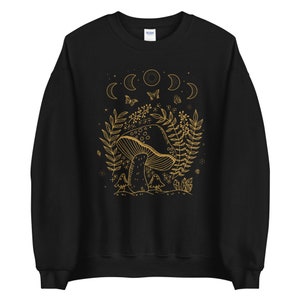 Op de afbeelding: Zwart sweatshirt met een gouden omtrek van een paddenstoel, maanfasen en vlinders. Het ontwerp is gecentreerd op de voorkant van het sweatshirt.
