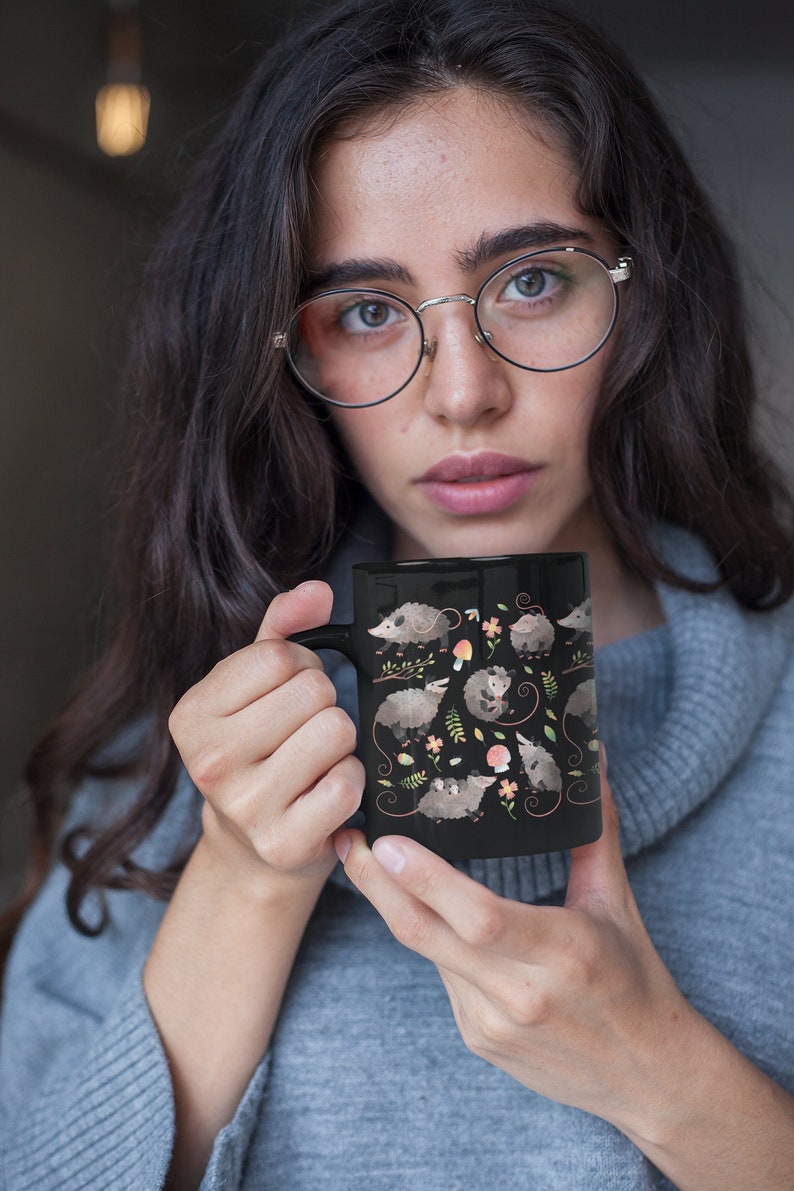 May include: A black ceramic mug with a white handle features a repeating pattern of opossums, mushrooms, and flowers.