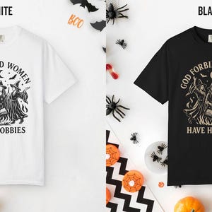 May include: A white t-shirt and a black t-shirt, each featuring a graphic print of three witches around a campfire with the text 'GOD FORBID WOMEN HAVE HOBBIES' arched above and below the illustration, surrounded by stars, bats, and a crescent moon.