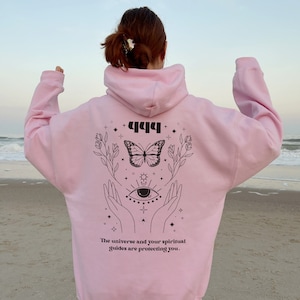 May include: Pink hoodie with a black graphic design featuring a butterfly, an eye, and the text "444 The universe and your spiritual guides are protecting you."