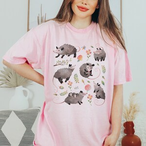 May include: A pink t-shirt with a graphic print of multiple opossums in a whimsical floral pattern. The opossums are depicted in various poses, including holding a baby opossum.