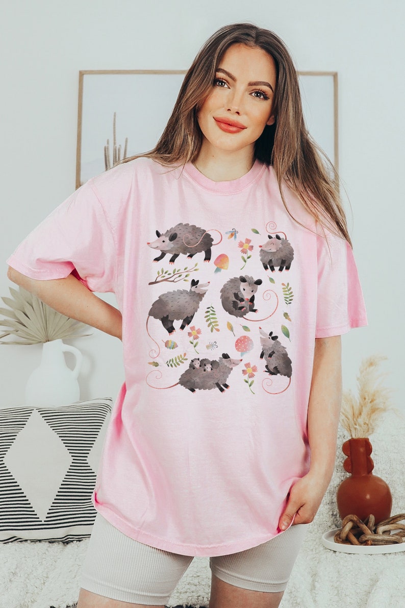 May include: A pink t-shirt with a graphic print of multiple opossums in a whimsical floral pattern. The opossums are depicted in various poses, including holding a baby opossum.