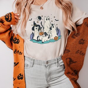 May include: A beige t-shirt with a graphic of five cats dressed in Halloween costumes. The cats are wearing witch hats, ghost costumes, and holding pumpkins. The graphic is surrounded by stars, bats, and a crescent moon.