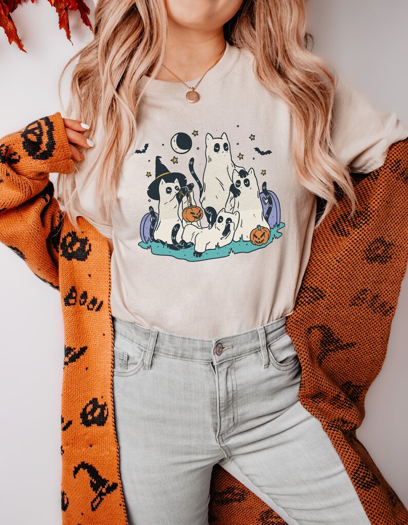 May include: A beige t-shirt with a graphic of five cats dressed in Halloween costumes. The cats are wearing witch hats, ghost costumes, and holding pumpkins. The graphic is surrounded by stars, bats, and a crescent moon.