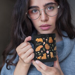 May include: A black ceramic mug with a white handle features a whimsical design of multiple capybaras in various poses, surrounded by flowers, leaves, and mushrooms. The capybaras are depicted in a light brown color.