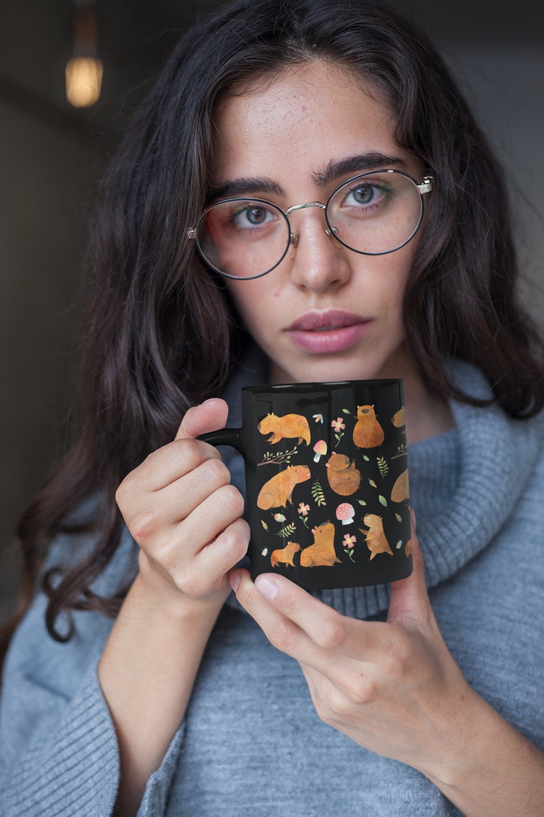 May include: A black ceramic mug with a white handle features a whimsical design of multiple capybaras in various poses, surrounded by flowers, leaves, and mushrooms. The capybaras are depicted in a light brown color.