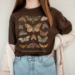 May include: A brown t-shirt featuring a print of various moths and butterflies. The text "We shall meet in the place where there is no darkness" is printed on the bottom of the shirt.