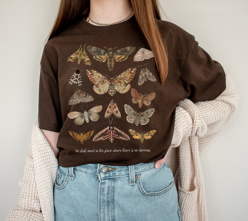 May include: A brown t-shirt featuring a print of various moths and butterflies. The text "We shall meet in the place where there is no darkness" is printed on the bottom of the shirt.