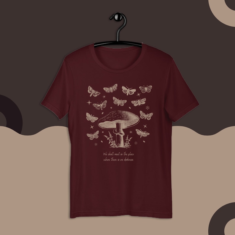 May include: A maroon t-shirt with a white graphic of a mushroom with a quote that reads "We shall meet in the place where there is no darkness". The graphic also includes a variety of moths and stars.