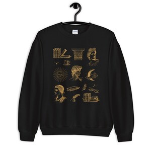 Puede incluir: Sudadera negra con un estampado dorado de varias ilustraciones que incluyen libros, un sol, una luna, una taza, una columna y algunas caras.