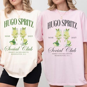 May include: Two white t-shirts with a graphic design of two glasses of Hugo Spritz, a cocktail, with the text "Hugo Spritz" and "Social Club" printed on the front. The text "MAR 2025" is printed below the glasses. The t-shirts are available in ivory and blossom colors.