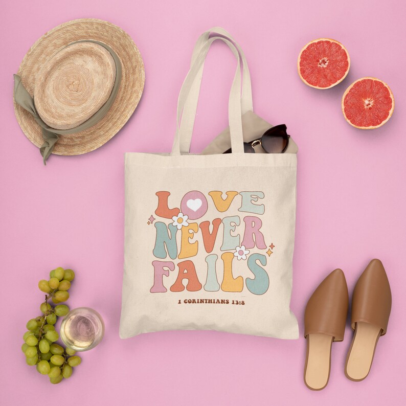 May include: A white canvas tote bag with the words "LOVE NEVER FAILS" in colorful retro-style lettering. The text is surrounded by small flowers and sparkles. The bottom of the bag has the text "1 Corinthians 13:8".