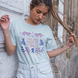 May include: A light blue t-shirt with a graphic design featuring pink tigers, snakes, leaves, and moons. The text on the shirt reads "You are made of magic".
