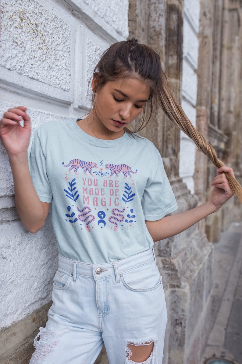 May include: A light blue t-shirt with a graphic design featuring pink tigers, snakes, leaves, and moons. The text on the shirt reads "You are made of magic".