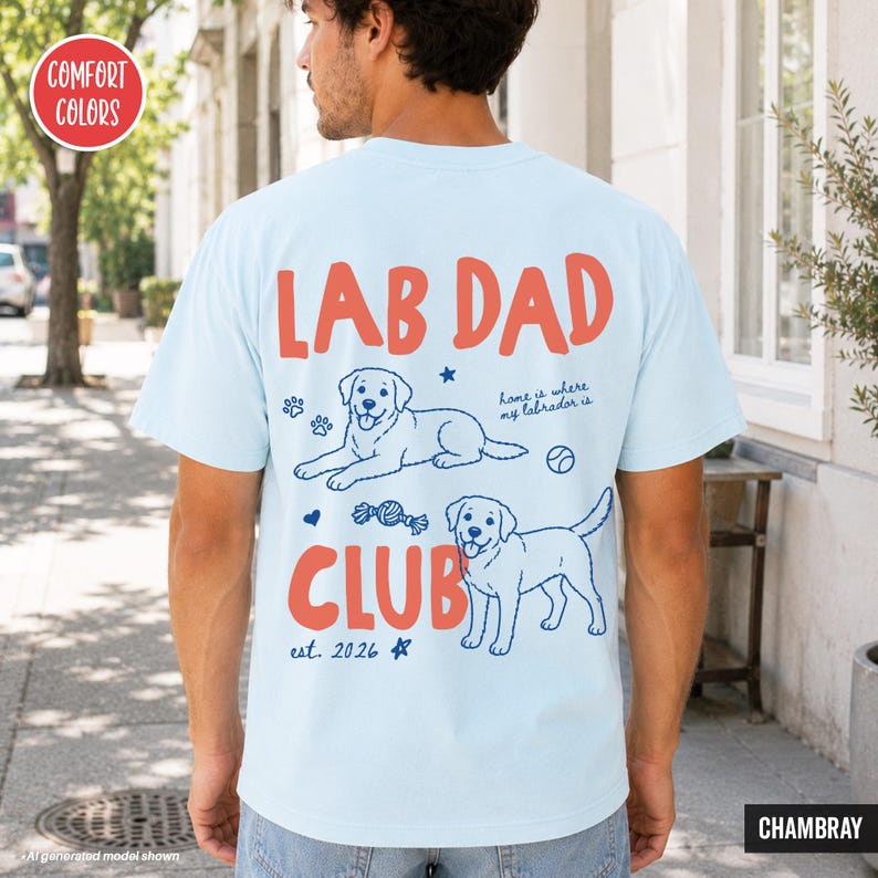 Puede incluir: Una camiseta azul claro con un gr&aacute;fico en la espalda que muestra el texto rojo LAB DAD CLUB. El dise&ntilde;o incluye dos perros Labrador en dibujo lineal, una pelota de tenis, un juguete de cuerda, huellas de patas y el texto home is where my labrador is y est. 2026.