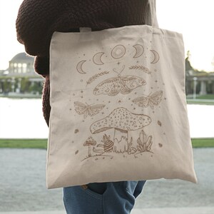 May include: A beige tote bag with a brown design of a mushroom, butterflies, and moons. The design is reminiscent of a mystical forest scene.